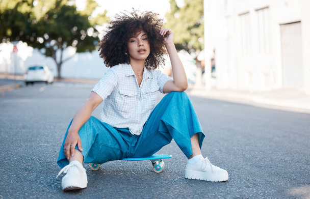 A young female mixed race woman skate sitting on a skateboard looking cool and confident with great style in a street outside. Hispanic hipster woman with curly afro hair style in a cool outfit - 写真・画像