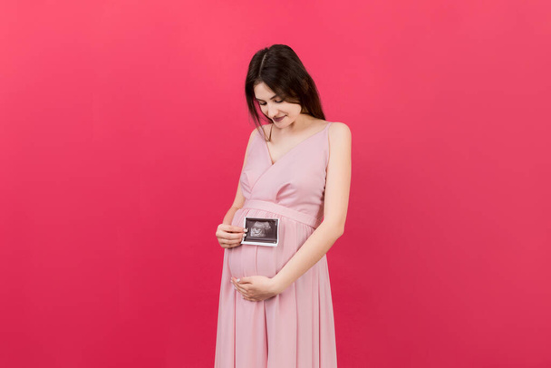 Schattige zwangere dame poseren met baby sonografie foto in de buurt van gekleurde achtergrond. Concept van zwangerschap, gynaecologie, medische test, gezondheid van de moeder. - Foto, afbeelding