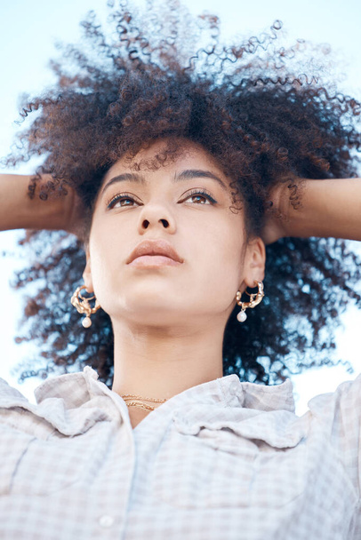 Closeup shot of a beautiful young mixed race female with her hands in her afro. Below shot of one african american woman looking thoughtful and contemplative while standing outside. Full of ideas. - Photo, image