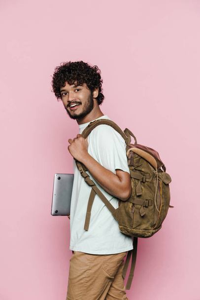 Young middle eastern man smiling while posing with laptop and backpack isolated over pink wall - 写真・画像