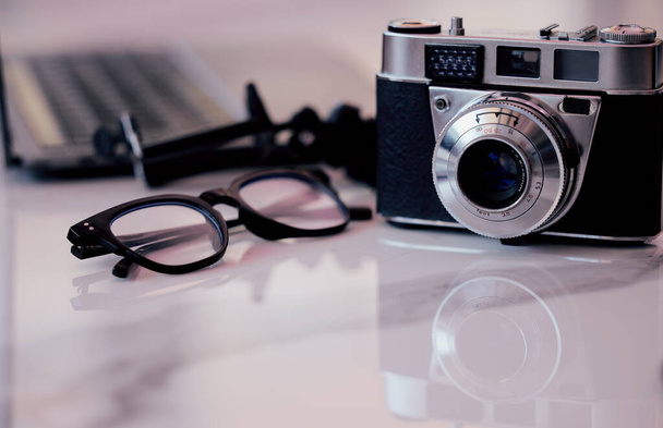 Closeup of a camera and glasses on a desk in an office. Blogger or photographers workstation at home of in a modern office. - Фото, изображение