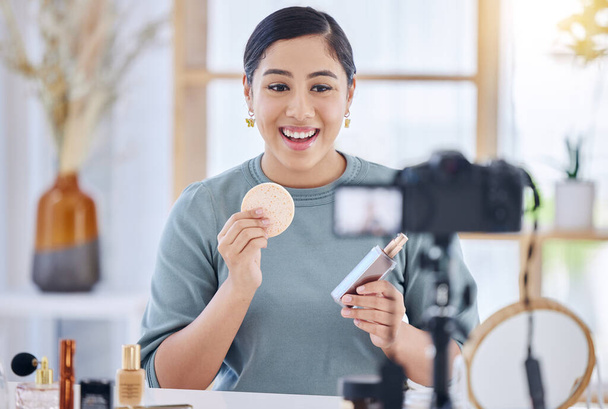 Happy young beauty blogger holding up a beauty product and sponge while doing a make-up tutorial and recording with her tripod. Influencer working as a brand ambassador for beauty products on social - Photo, Image