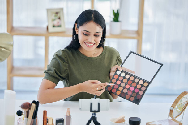 One beautiful mixed race creative woman sitting alone in a studio and using her cellphone to vlog while showing her eyeshadow palette. Happy hispanic makeup artist filming a tutorial for the internet. - Fotografie, Obrázek
