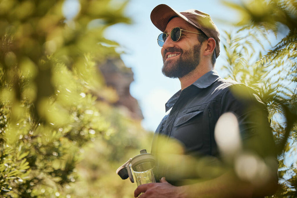 Adventurous fit man wearing glasses and a cap while out hiking or exploring the woods. Handsome man with beard smiling while exploring nature in the forest. - Photo, Image