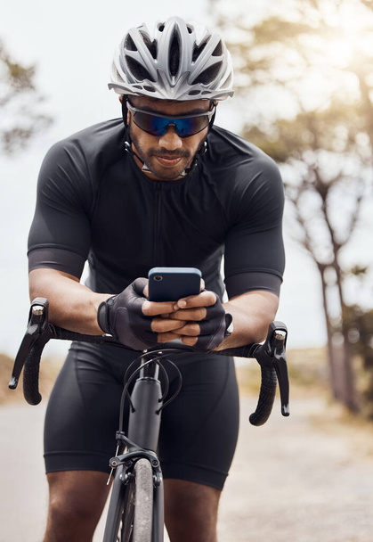 Male cyclist wearing helmet and glasses while using his smartphone and sitting on his bike. Fit sportsman using mobile phone while cycling outdoors. - 写真・画像