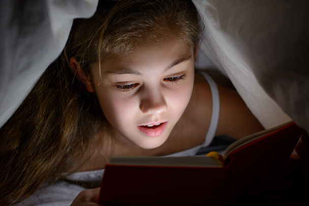 Impressed little girl under the blanket with a book and a flashlight reading horrors before go to bed. Small female child covered with blanket and enjoy reading scary stories in her bed at night - Fotó, kép