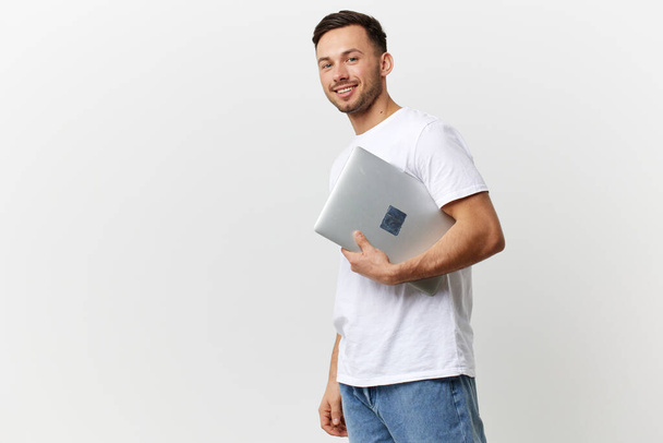 Lächelnd hält ein braun gebrannter, gutaussehender Mann im einfachen T-Shirt einen Laptop in die Kamera und posiert isoliert auf weißem Studiohintergrund. Kopierraum Banner-Attrappe. Elektronisches Reparaturkonzept - Foto, Bild