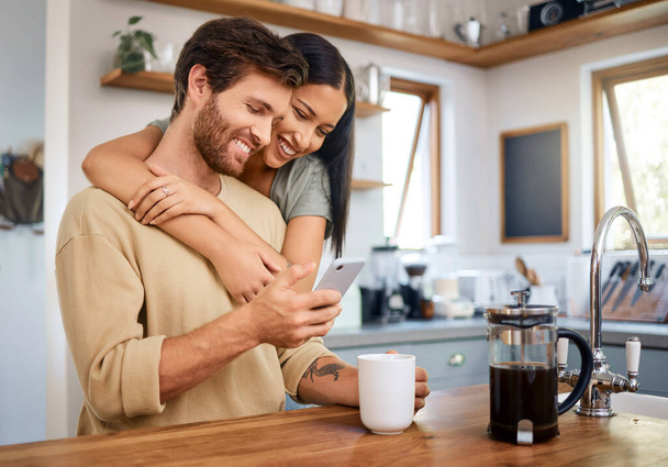 Happy young interracial couple being loving and affectionate at home. Young man using his smartphone and holds coffee cup while his girlfriend embraces him from behind. Browsing social media or - Zdjęcie, obraz