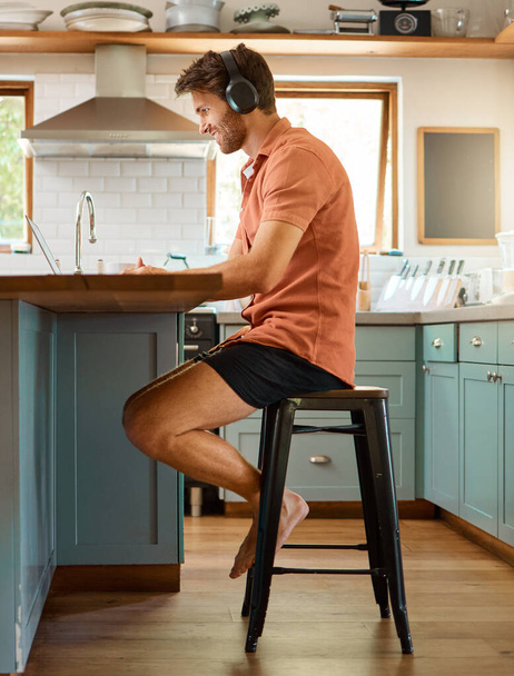 Fullbody of a young happy caucasian businessman wearing headphones and using a laptop at home alone. One joyful male businessperson smiling while working in the kitchen at home. - Foto, Imagen