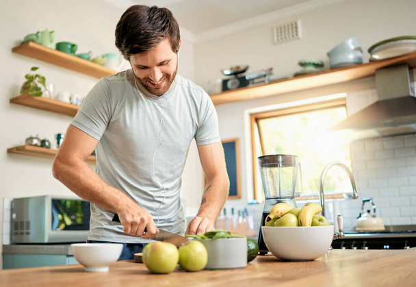 One fit young caucasian man cutting ingredients with knife to make healthy green detox smoothie while wearing earphones in kitchen at home. Guy having fresh fruit juice to cleanse and provide energy - 写真・画像
