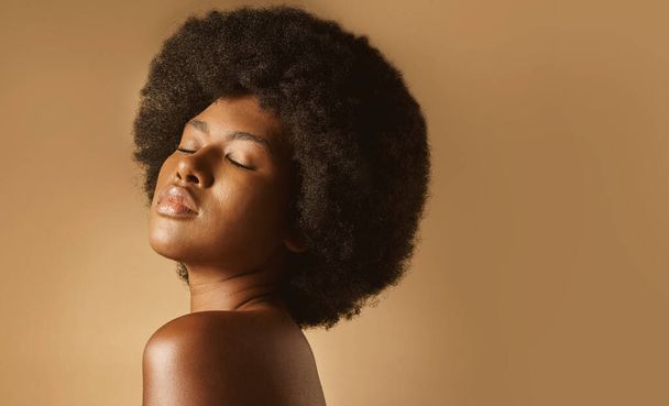 Selbstbewusste Afroamerikanerin mit natürlichem Afro, die vor braunem Studiohintergrund posiert. Junge schöne schwarze Model sieht trendy, kraftvoll und sinnlich mit glatt glühender Haut. - Foto, Bild