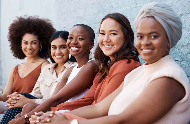 Group of diverse happy businesswomen sitting in a line with coworkers against a wall outside in the city. Businesspeople smiling while sitting outdoors with colleagues. - Fotó, kép