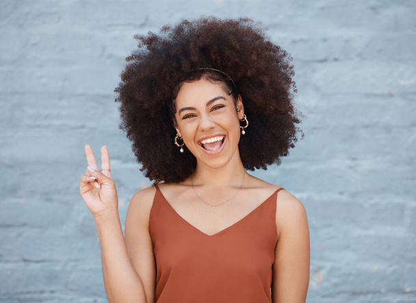 Eine junge gemischte Geschäftsfrau mit einem Afro steht draußen vor einer grauen Wand und gestikuliert mit ihrer Hand das Friedenszeichen. Glückliche und selbstbewusste weibliche Führungskräfte sehen positiv und erfolgreich aus. - Foto, Bild