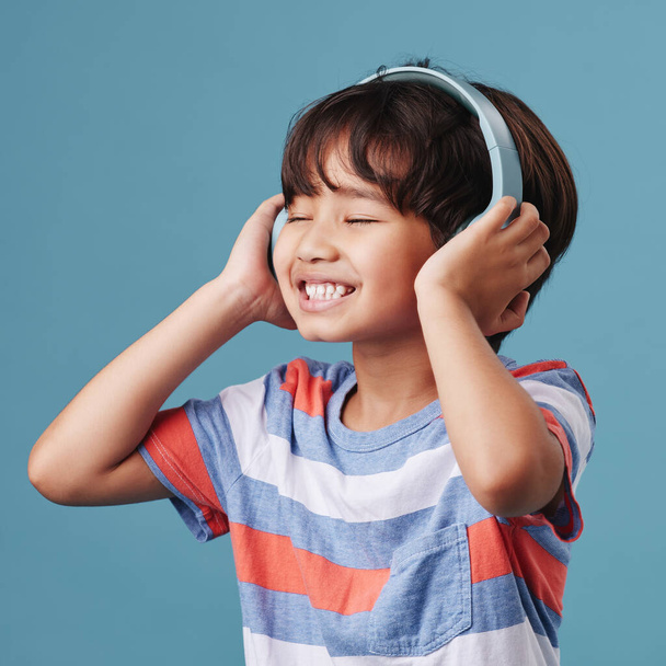 A cute young asian boy enjoying listening to music from his headphones. Adorable Chinese kid feeling the magic of music while posing against a blue studio background. - Foto, imagen