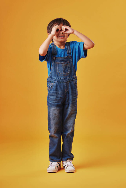 Niño curioso mirando a través de los dedos en forma de prismáticos mientras usa ropa casual sobre un fondo de estudio amarillo. - Foto, imagen