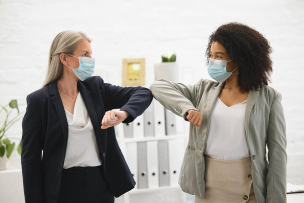 Two diverse colleagues talking and greeting each other with their elbows while wearing masks to protect from infection of a virus. Businesswomen joining their elbows to greet in an office at work. - Foto, imagen