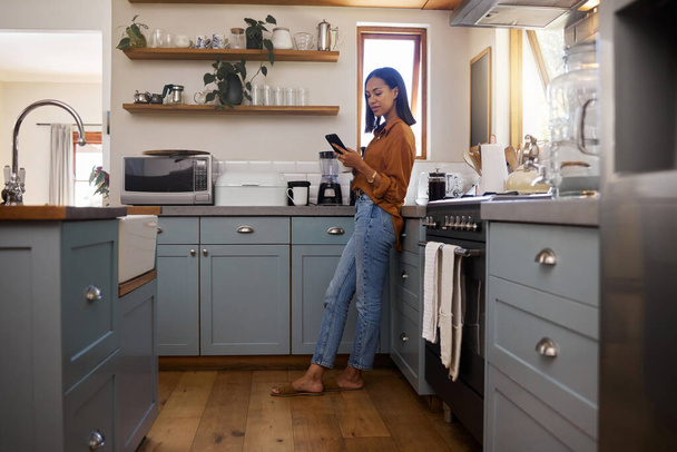 Young mixed race woman drinking a cup of coffee while using her phone alone in the morning in the kitchen. One hispanic female enjoying a cup of tea while using social media on her phone at home. - Фото, изображение