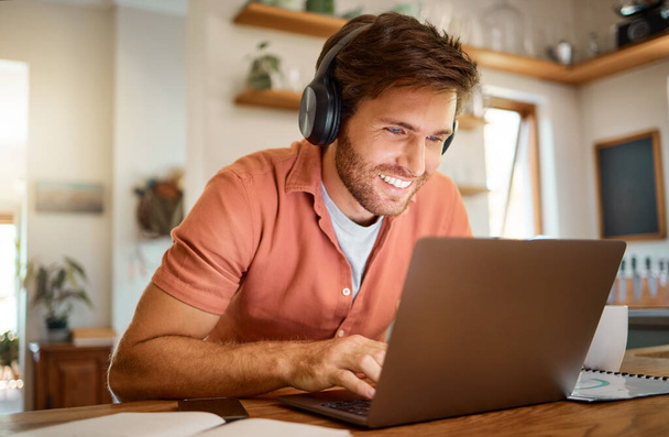 Young cheerful caucasian businessman wearing headphones and listening to music while using a laptop at home alone. One content male businessperson typing on a laptop while working in the kitchen at - Photo, Image