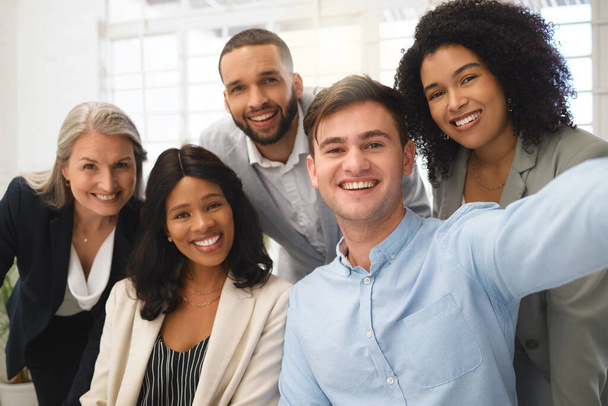 Group of cheerful diverse businesspeople taking a selfie together at work. Happy young caucasian businessman taking a photo with his content colleagues in an office. Coworkers bonding at work. - Fotografie, Obrázek