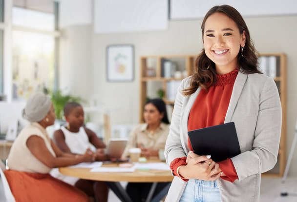 Smiling asian business woman holding a digital tablet while colleagues sit behind her in office. Ambitious and happy professional standing after browsing schedule on technology before leading meeting. - Photo, Image