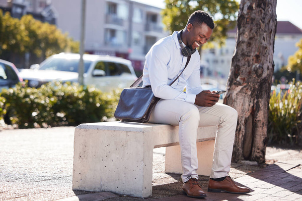 Un hombre afroamericano feliz usando auriculares alrededor de su cuello y mensajes de texto en un teléfono mientras estaba en la ciudad en su viaje diario. Un guapo hombre negro sonriendo mientras revisa su feed en las redes sociales. - Foto, Imagen