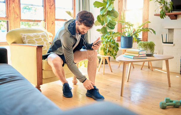 One fit young caucasian man getting dressed for exercise workout while listening to music with earphones on a cellphone at home. Guy sitting on a chair putting on sports shoe sneakers while getting - Foto, Imagem