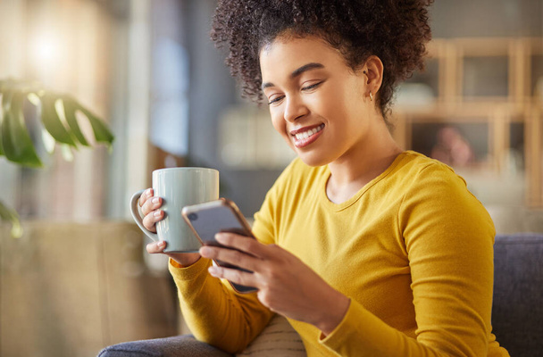 Szczęśliwa mieszanka kobiet rasy pije filiżankę kawy i wpisuje wiadomość na telefonie w domu. Jedna zadowolona latynoska kobieta z kręconym afro za pomocą mediów społecznościowych w telefonie komórkowym podczas relaksu na kanapie w - Zdjęcie, obraz