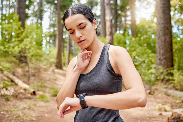 One beautiful young fit and athletic woman checking her pulse while exercising outdoors. An attractive mixed race female checking her heart rate on her smart watch during a workout in nature. - Valokuva, kuva