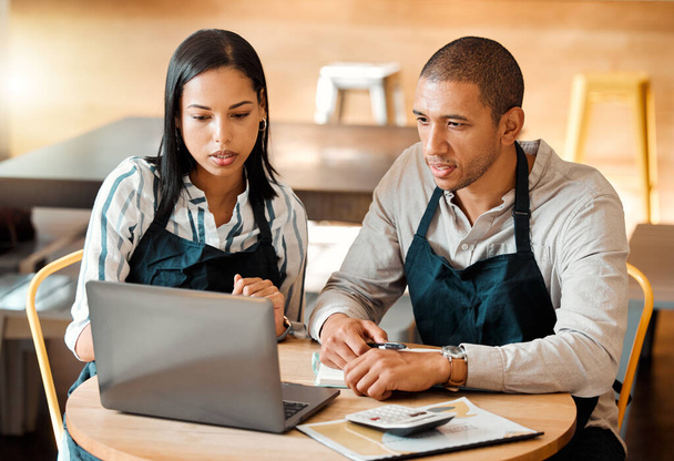 Two young mixed race employees wearing aprons sitting at a table in a coffee shop using a laptop and calculator at work. Young male and female hispanic colleagues brainstorming as a team because alone - Φωτογραφία, εικόνα