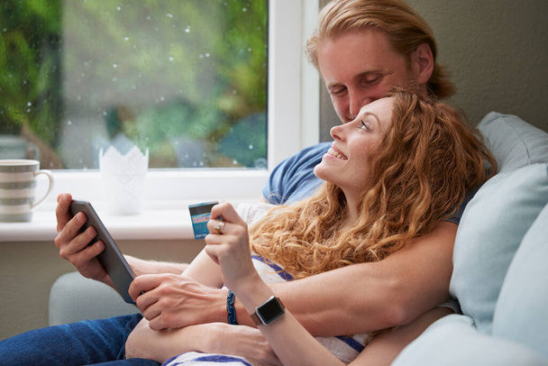 Ένα νεαρό ευτυχισμένο ζευγάρι που αγκαλιάζεται ενώ χρησιμοποιεί ένα ψηφιακό tablet για να κάνει online πληρωμές με κάρτες. - Φωτογραφία, εικόνα