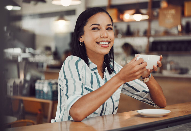 Szczęśliwy młody uśmiechnięty mieszane rasy kobieta w casual strój pije gorący napój siedząc samotnie przy stole w przytulnej kawiarni. Piękna kobieta wygląda szczęśliwy i przemyślany picia kawy i korzystających z niej - Zdjęcie, obraz