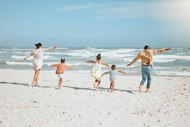 Playful mixed race family with three children running on the beach with arms outstretched. A big family spending time and enjoying holiday by the sea. - 写真・画像