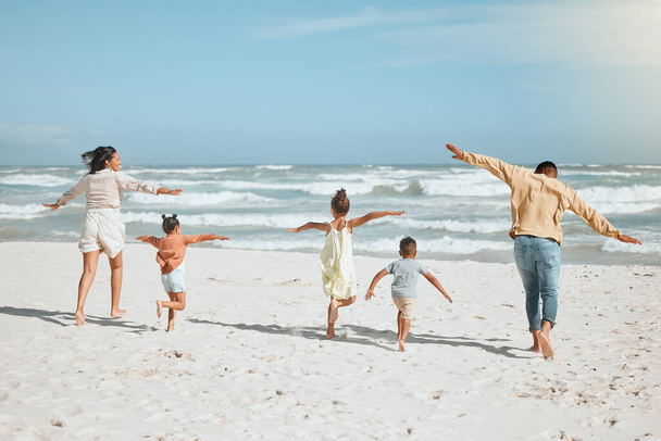 Zadní pohled na hravou smíšenou závodní rodinu se třemi dětmi běžícími na pláži s nataženýma rukama. Velká rodina trávit čas a užívat si dovolenou u moře. - Fotografie, Obrázek