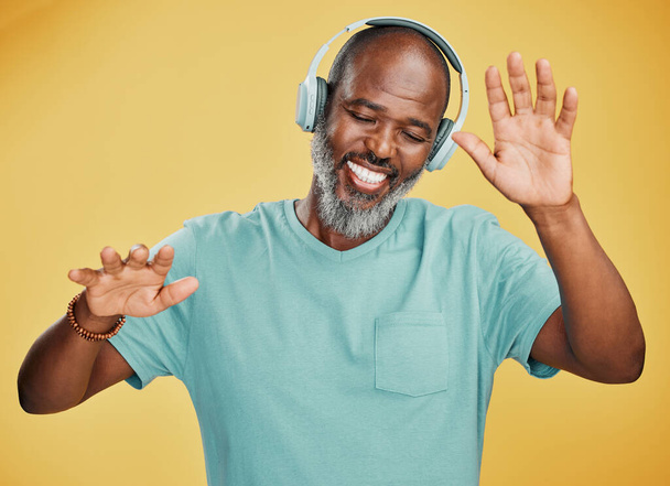 Szczęśliwy dojrzały Afrykanin tańczący słuchając muzyki ze słuchawkami na żółtym tle studia. Beztroski starszy czarny mężczyzna z szarą brodą świętujący i cieszący się ulubionymi piosenkami. - Zdjęcie, obraz