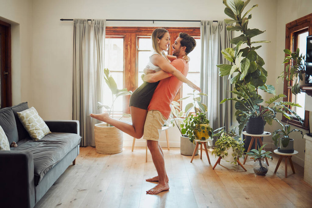 Happy young boyfriend holding girlfriend in arms as he lifts her up while they look into each others eyes and share intimate moment. Romantic young couple hugging and enjoying passionate dance at home - Photo, image