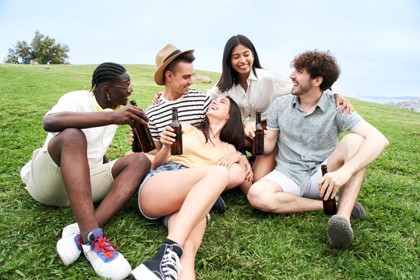 Retrato al aire libre de grupo de amigos brindando con botellas de cerveza sentados en la hierba celebrando juntos divertirse. Vibraciones veraniegas. Foto de alta calidad - Foto, imagen
