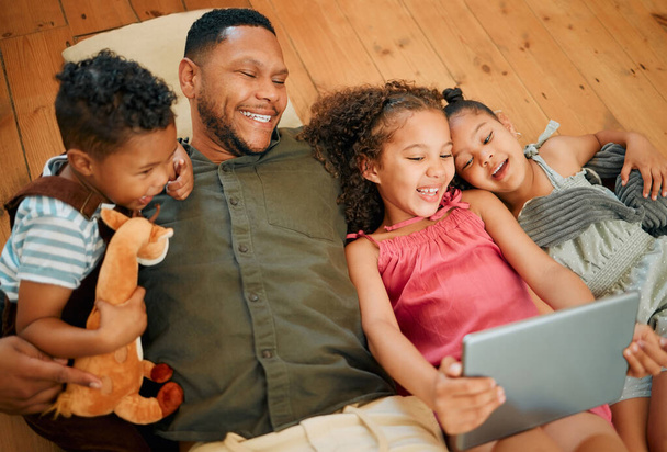 A happy mixed race family of four relaxing and lying on the lounge floor together. Loving black single parent bonding with his kids while using a digital tablet to watch movies. - Valokuva, kuva