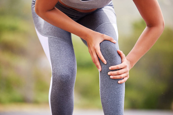 Active woman holding her leg in pain while exercising outdoors. Closeup of an athlete suffering with a painful knee injury, causing discomfort and strain. Muscle sprain from a fractured joint. - Фото, изображение