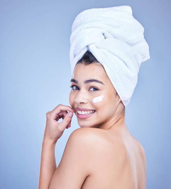 Studio Portrait of a beautiful smiling mixed race woman applying cream to her face. Hispanic model with glowing skin and wet hair against a blue copyspace background. - 写真・画像
