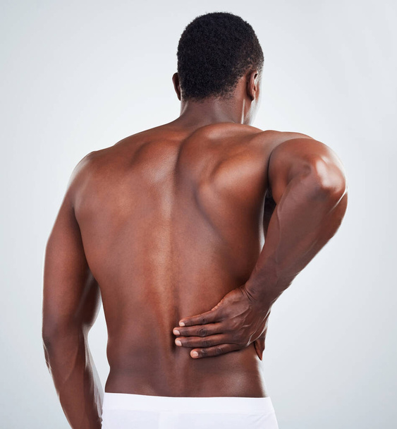 One unknown muscular fitness model experiencing back pain from poor posture while exercising. Black topless athlete with backache while isolated on grey copyspace in a studio. - Photo, Image
