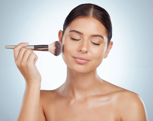 Beautiful woman holding and using cosmetic makeup brush to apply blush makeup while posing with copyspace. Caucasian model isolated against grey studio background. Getting ready after skincare routine - Photo, Image
