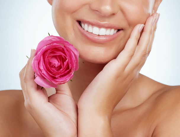 Посміхнена незнайома жінка, що тримає рожеву троянду, притулившись до обличчя. Кавказька модель, ізольована від сірого студійного фону з гладкою блискучою шкірою і делікатними здоровими звичками догляду за шкірою. - Фото, зображення