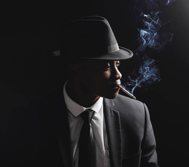 Ένας νεαρός Αφροαμερικάνος που δείχνει σοβαρός καπνίζοντας ένα τσιγάρο πάνω σε μαύρο φόντο. Αφρικανός που μοιάζει με τον εαυτό του βαθιά στη σκέψη φορώντας ένα μαύρο επίσημο κοστούμι και ένα καπέλο με απομονωμένη - Φωτογραφία, εικόνα