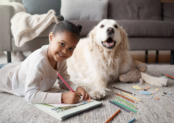 Sweet little mixed race child doing her homework while lying on the living room carpet with her puppy. Child colouring while bonding with her emotional support rescue dog. - Fotoğraf, Görsel