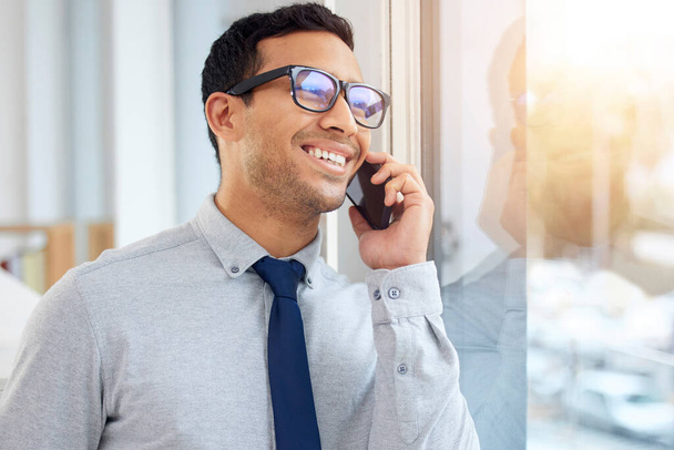 Young content mixed race businessman on a call using a phone and standing in an office at work. One hispanic male businessperson smiling while talking on the phone standing and thinking at a window in - Фото, зображення