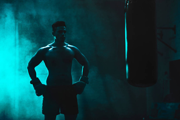 Silhouette of an unrecognizable boxer man sparring with a punching bag in a dark gym. Male athlete working out in preparation for his upcoming fight. Young martial artist training and practicing. - Fotoğraf, Görsel