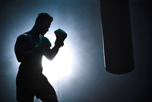 Silueta nerozpoznatelného boxera zápasícího s boxovacím pytlem v tmavé tělocvičně. Mužský atlet cvičí v přípravě na svůj nadcházející zápas. Mladý bojový umělec trénink a trénink. - Fotografie, Obrázek