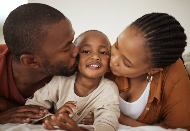 Porträt eines entzückenden kleinen afrikanisch-amerikanischen Jungen, der zu Hause zwischen Mutter und Vater auf einem Bett liegt. Liebevolle Eltern küssen ihren kleinen Sohn auf die Wangen. - Foto, Bild