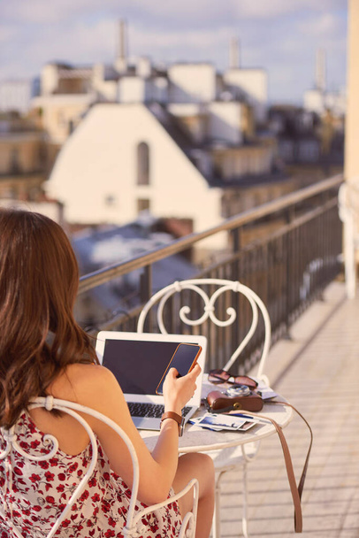 Shot of a woman using a smartphone and laptop while having breakfast on the balcony of an apartment overlooking Paris, France. - Foto, Imagem