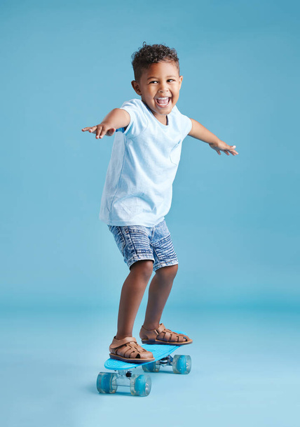 Schattige kleine latino jongen kijkt opgewonden terwijl balanceren met zijn armen uitgestrekt en rijden zijn skateboard tegen een blauwe studio achtergrond. - Foto, afbeelding